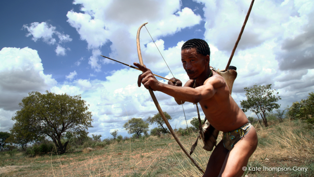A person wearing traditional clothing aims a bow and arrow in a grassy, open landscape with scattered trees under a partly cloudy sky. The individual is in a dynamic pose, demonstrating focus and readiness to shoot. © Kate Thompson-Gorry.