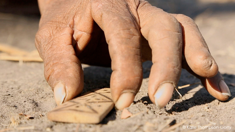 A close-up of a person's hand reaching to pick up a small, rectangular, carved object on the ground. The hand appears to be dusty, suggesting an outdoor setting. The object has geometric designs on its surface. Sunlight illuminates the scene.