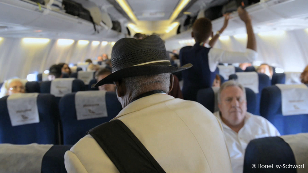 An elderly man wearing a white jacket and black hat sits in an airplane aisle seat, facing forward. Passengers are seated around him, and a flight attendant assists with luggage. The airplane cabin is well-lit with overhead compartments visible. © Lionel Isy-Schwart.