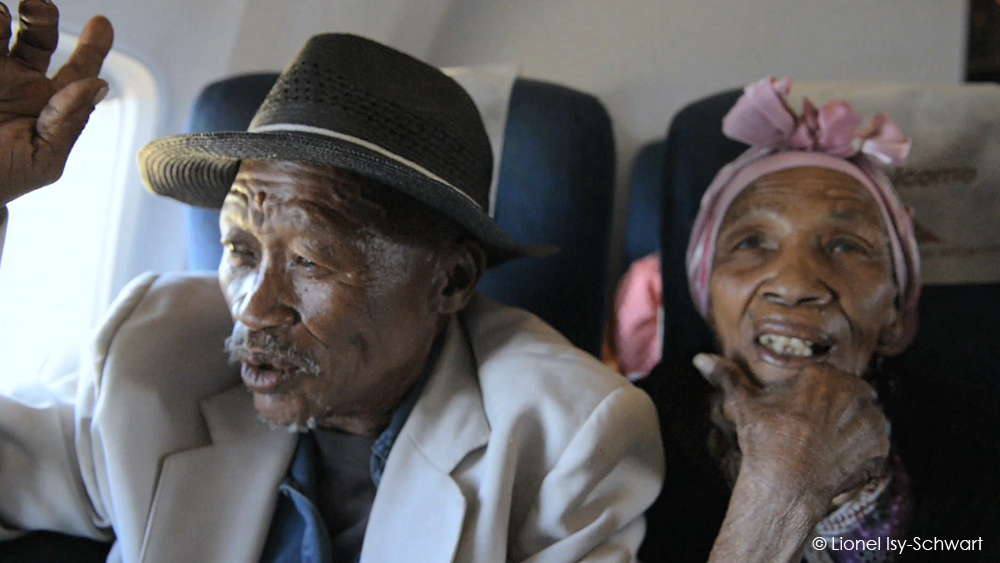 Two elderly individuals sit in an airplane. The person on the left, wearing a fedora and light-colored blazer, appears to be speaking. The person on the right, adorned with a pink headscarf and dark clothing, smiles with a thoughtful expression. They are seated next to each other.