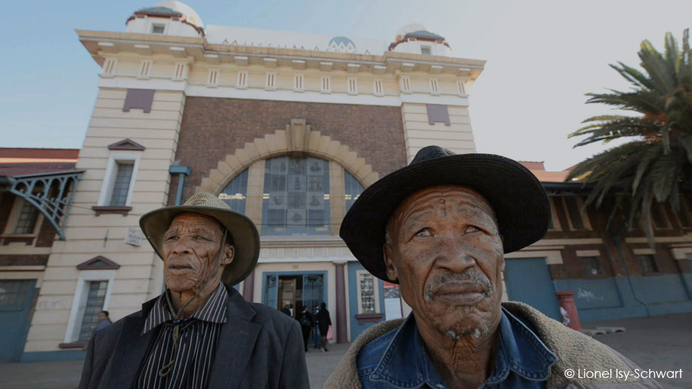 Two elderly men wearing hats stand in front of a historic building with large arched windows and ornate architecture. The building's facade features a mix of brick and decorated stone. A few people are visible near the entrance. Palm trees are seen on the right. © Lionel Isy-Schwart.