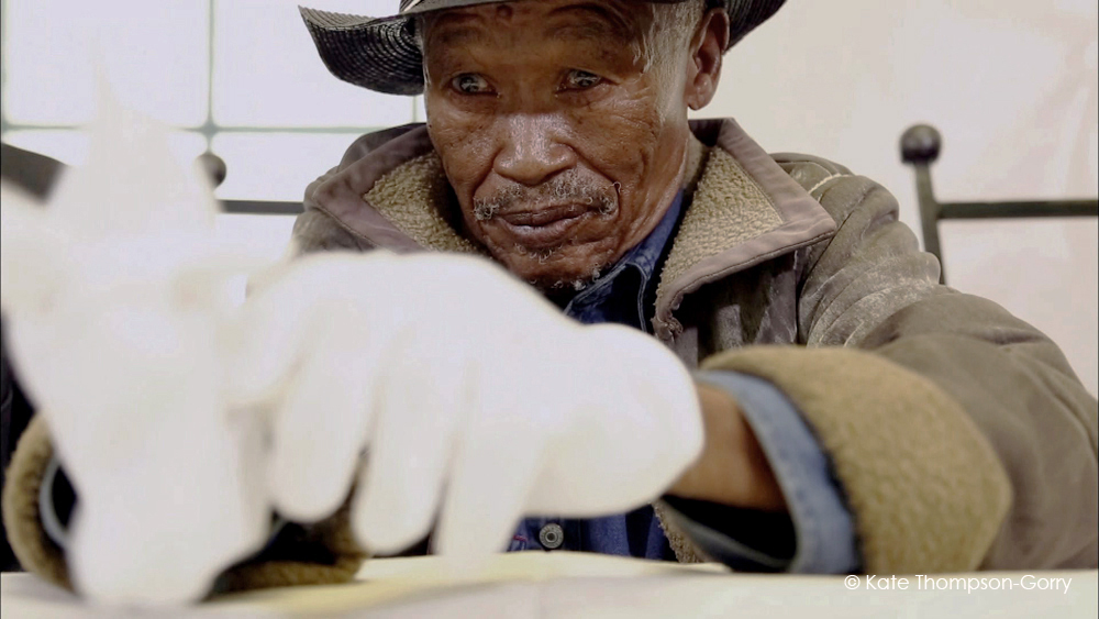 An elderly man with a weathered face and a mustache, wearing a dark hat and a brown jacket with a shearling collar, looks intently at something in front of him. His hands are outstretched, wearing white gloves. © Kate Thompson-Gorry is noted in the corner.